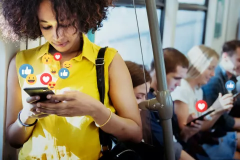 Eine Frau im Vordergrund schaut sich einen Social-Media-Post an und interagiert mit ihm. Im Hintergrund sind weitere Menschen, die in Social-Media vertieft sind und Likes und Herzen setzen.